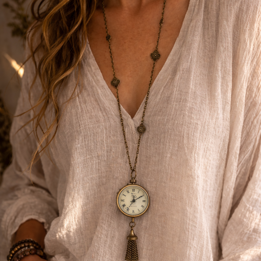 Person wearing a long necklace with a clock pendant, dressed in a light-colored outfit.